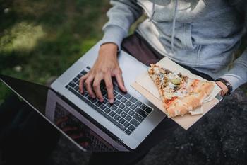 Image of a college student using a laptop with a slice of pizza in their hand